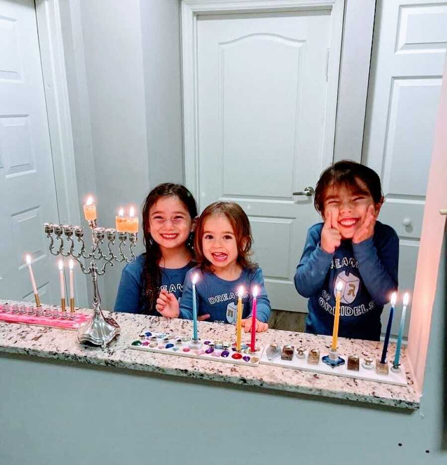Mom takes a photo of her three daughters smiling hard while lighting candles in the menorah for Hanukkah