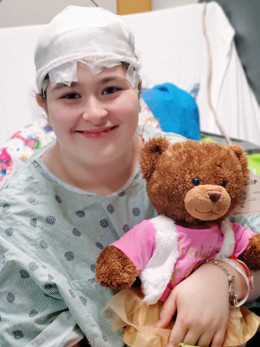 Young girl with epilepsy sits in a hospital bed with bandages wrapped around her head while holding a teddy bear
