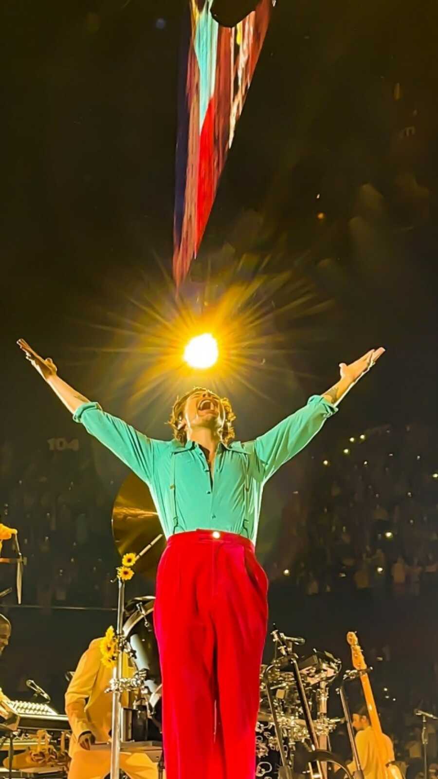 Harry Styles performing at a concert with a beautiful light above him while he wears red pants, and a green button down