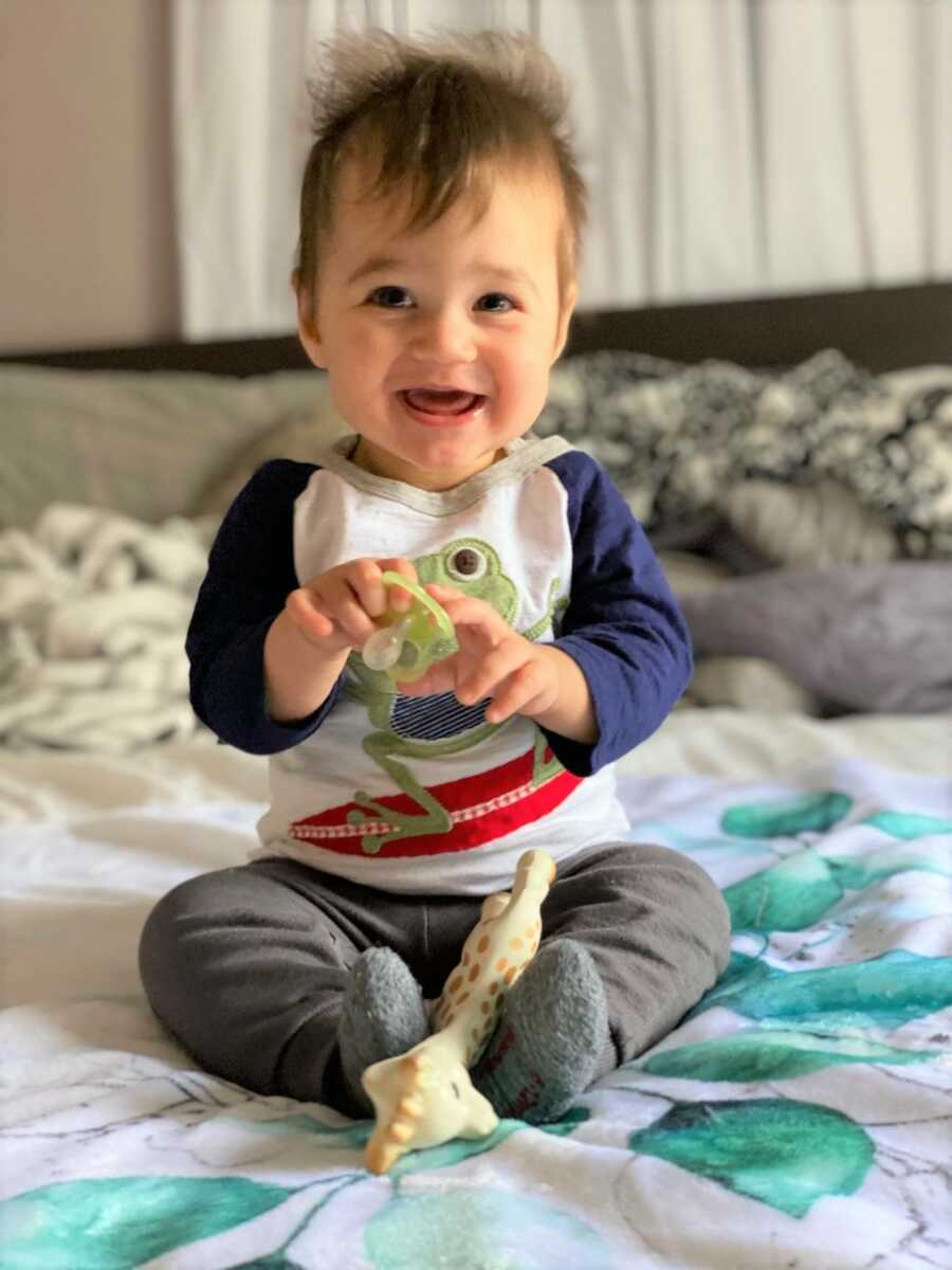 Sami sits and smiles at the camera while playing with his pacifier. 