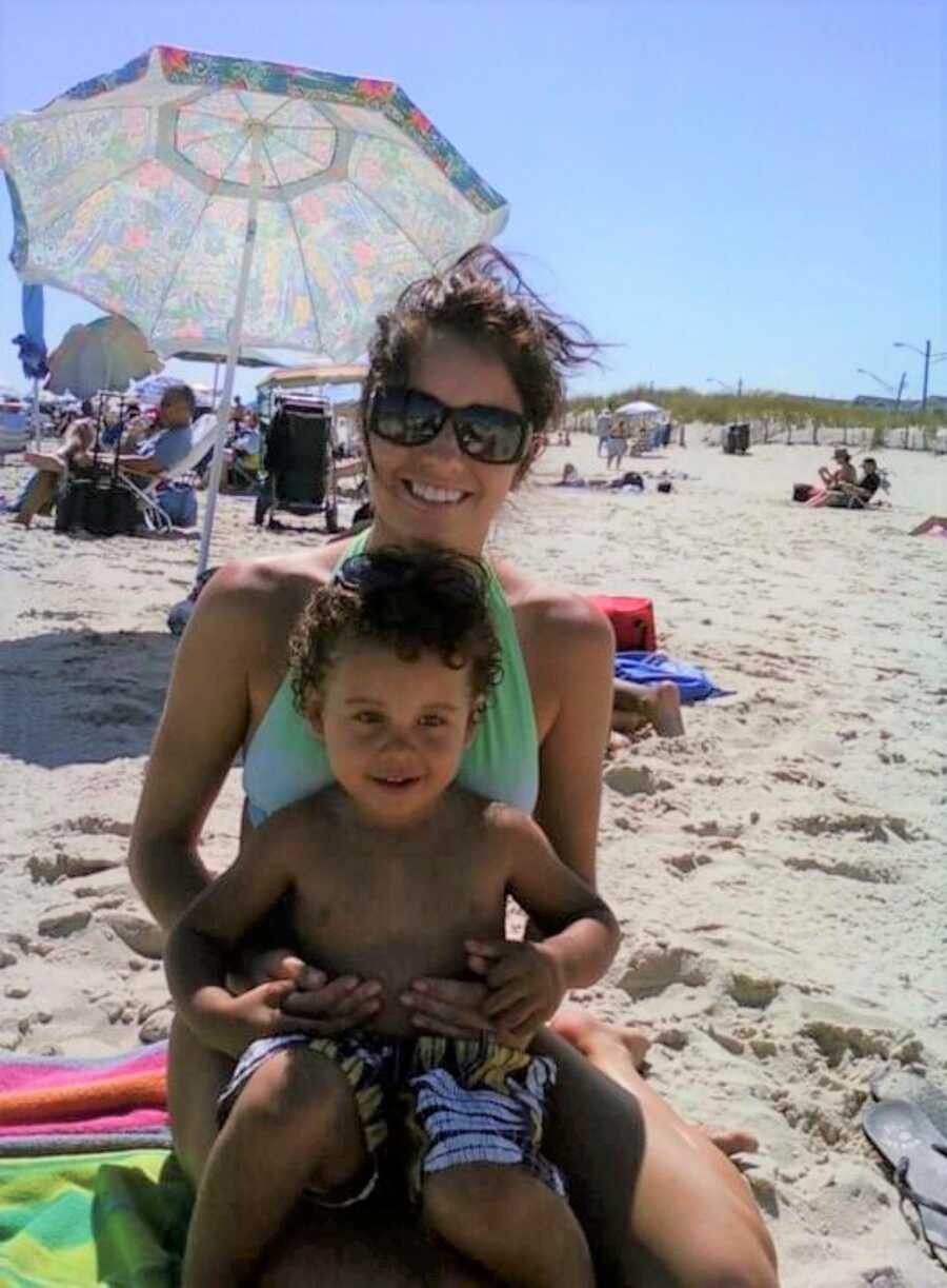 Mom and toddler son sit on the sand over a towel at the beach with an umbrella in the background