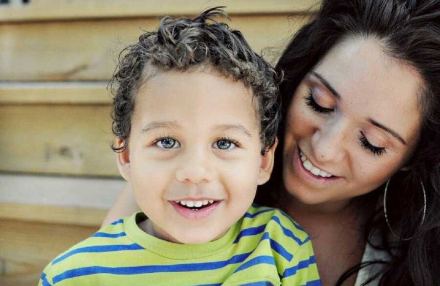 toddle son with bright green eyes looks straight at the camera while mom hugs him sitting on the stairs 