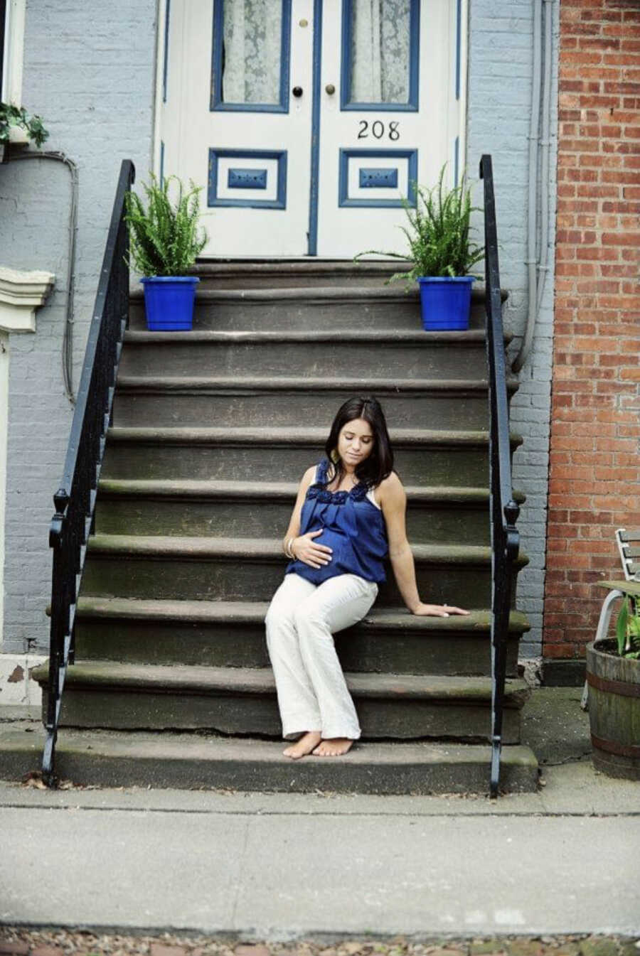 pregnant woman sits on stairs outside of an apartment while holding pregnant belly wearing blue shirt with blue plant pots in the background next to brick wall