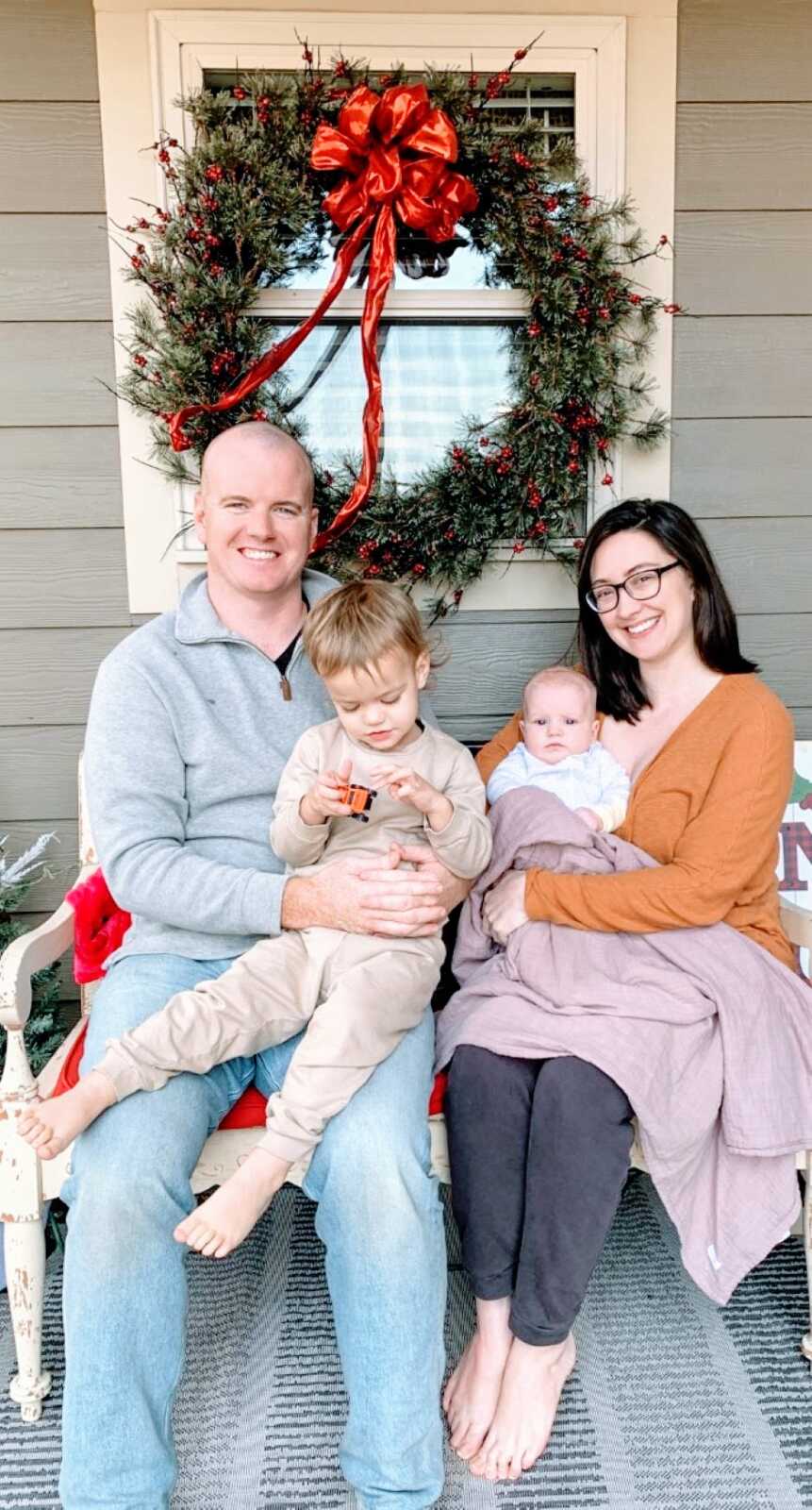 Family of four take a photo on their front porch with a Christmas wreath behind them