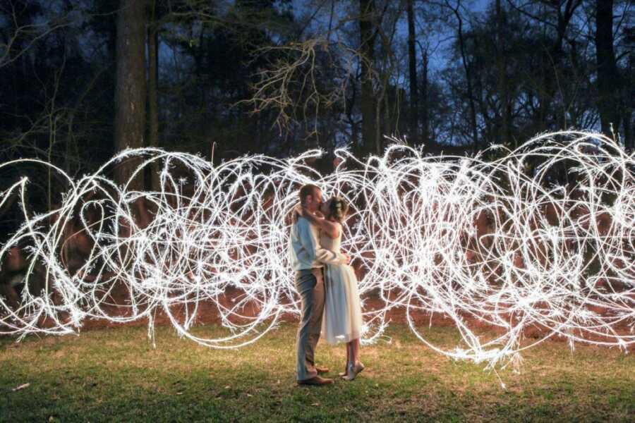 Newlyweds kiss at their wedding while guests make shapes with sparklers behind them