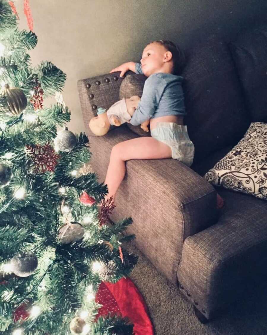 little boy with his teddy bear sitting on the arm of the couch looking at the christmas tree