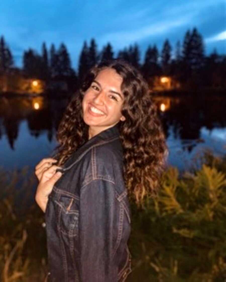 Young woman stands in front of a lake at night while wearing a denim jacket