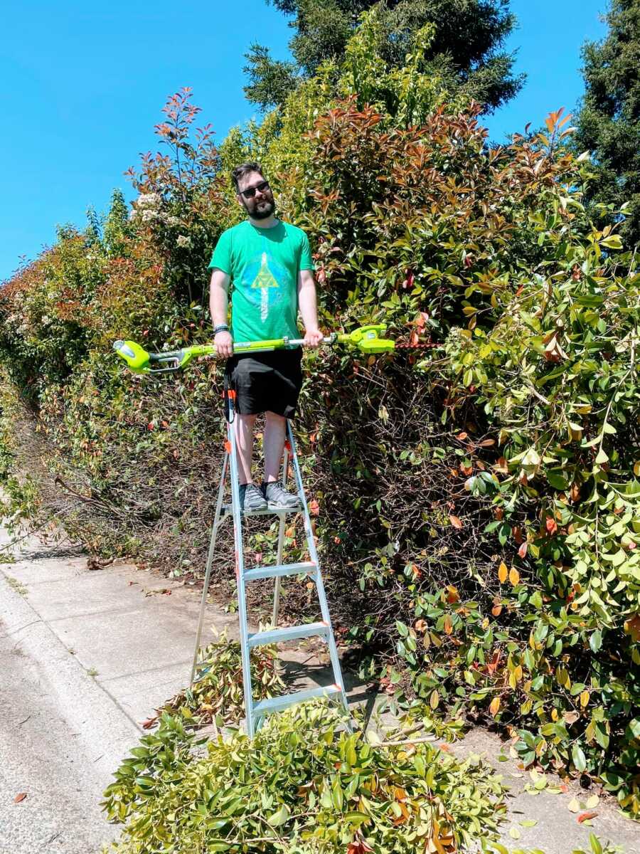 Man smiles on a ladder while trimming hedges on the sidewalk