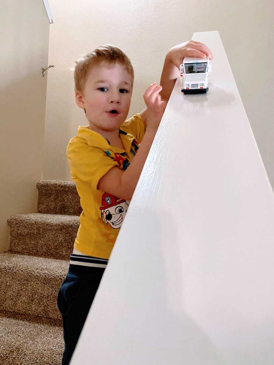 Little boy in a Paw Patrol shirt plays cars on the banister of a staircase