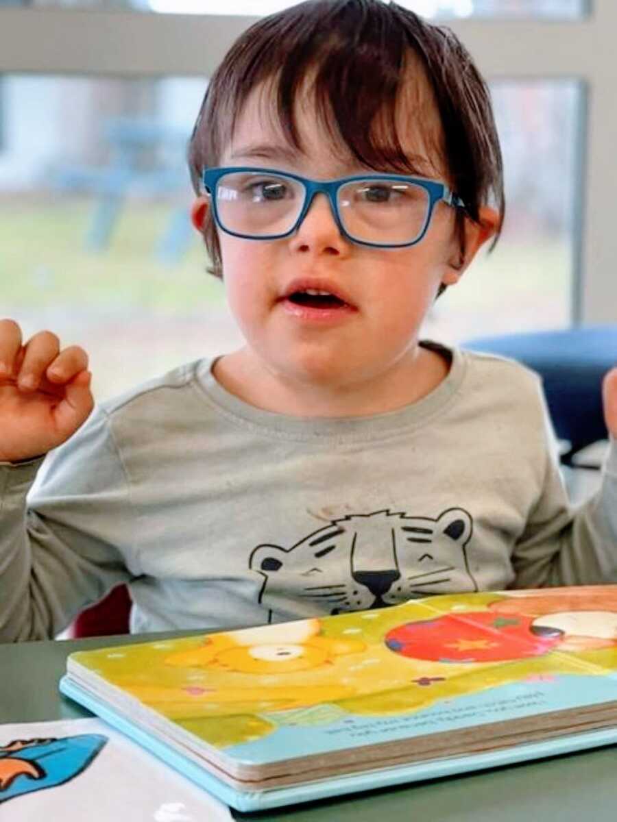 Little boy with down syndrome poses for the camera while wearing blue glasses and reading a book