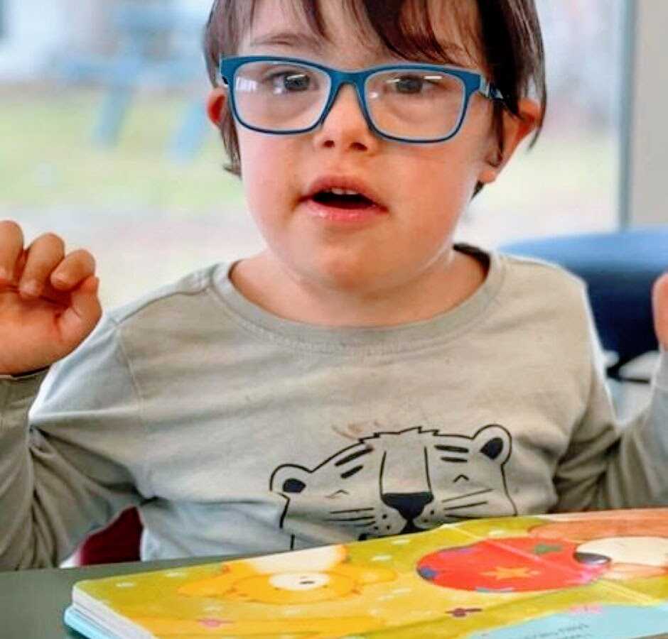 Little boy with down syndrome poses for the camera while wearing blue glasses and reading a book