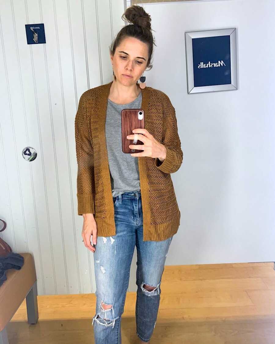 women in fitting room takes mirror selfie wearing a cardigan and jeans
