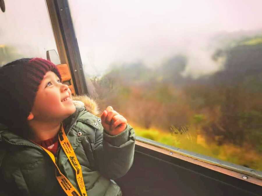 young boy in winter gear faces towards window and looks out at greenery