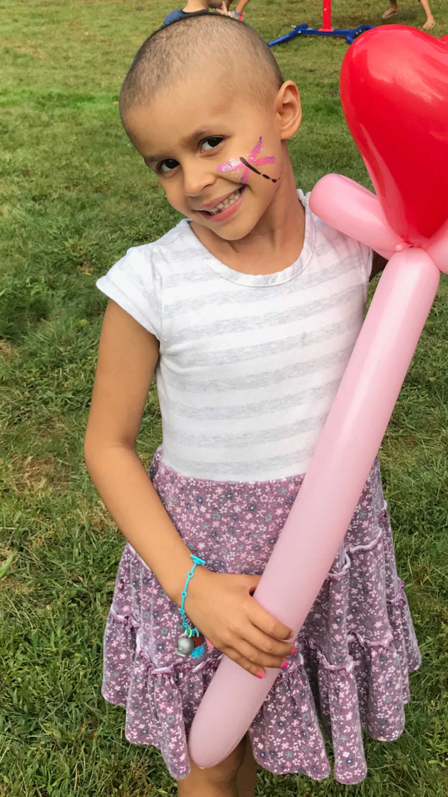 young girl with childhood cancer smiling at camera with face paint on and a balloon animal