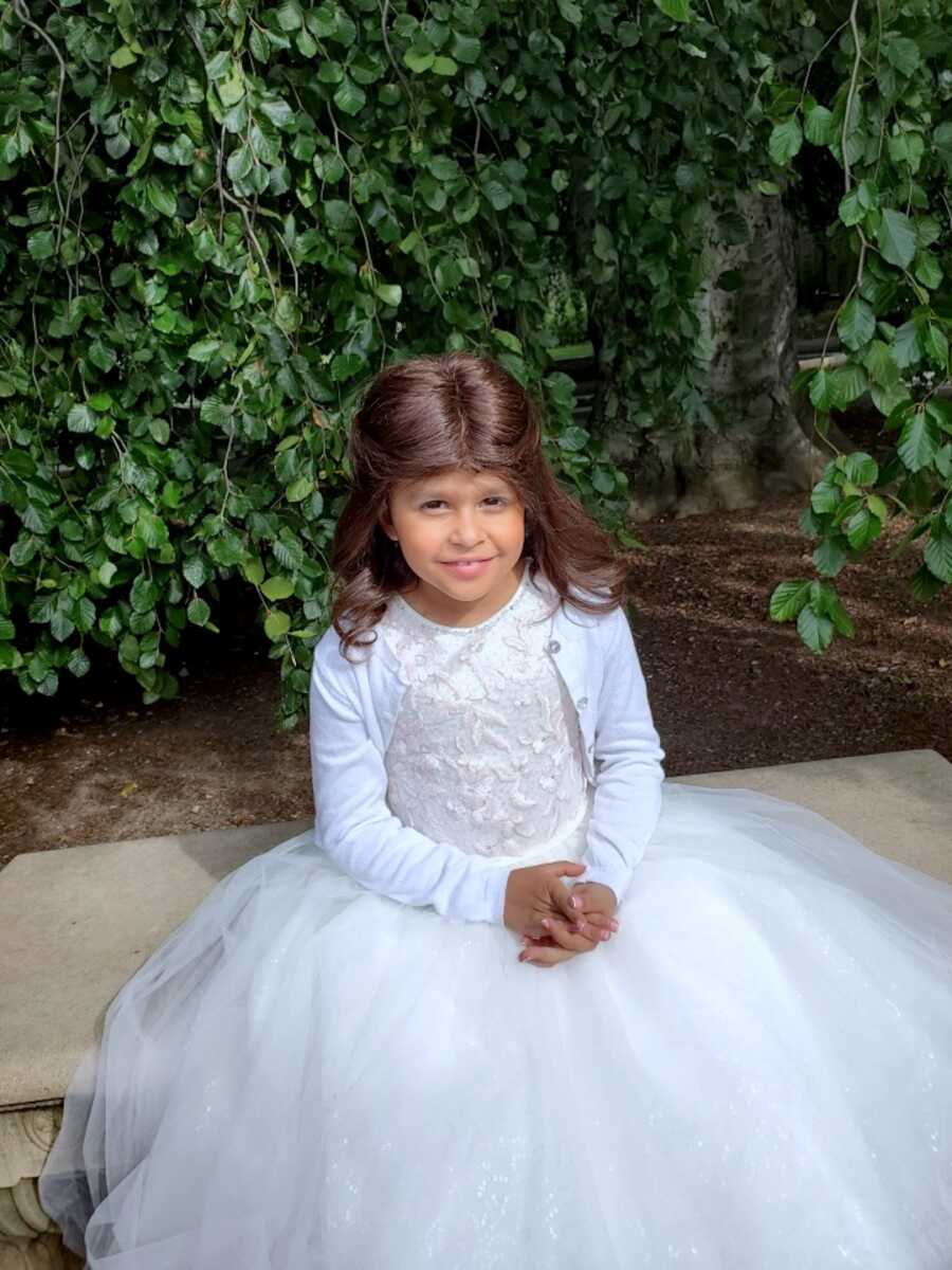 young girl sitting in front of bushes in a white dress and cardigan