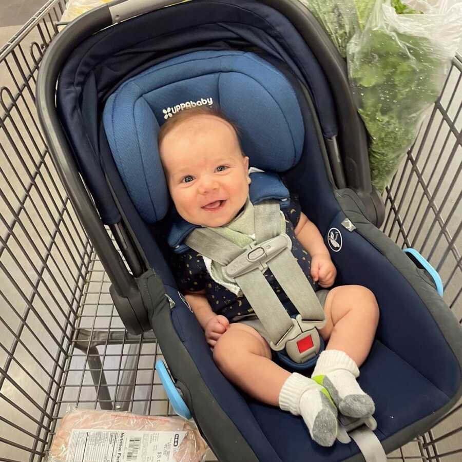 Baby in car seat sits in grocery cart and smiles at strangers.
