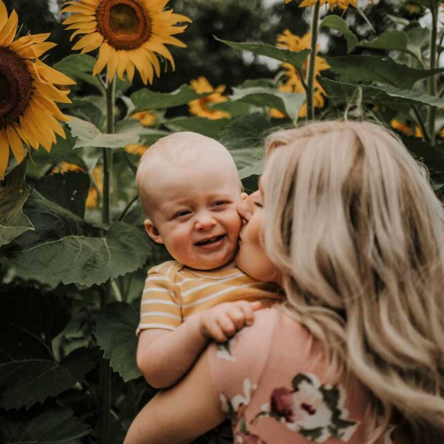 Mom kisses baby boy in sunflower patch.