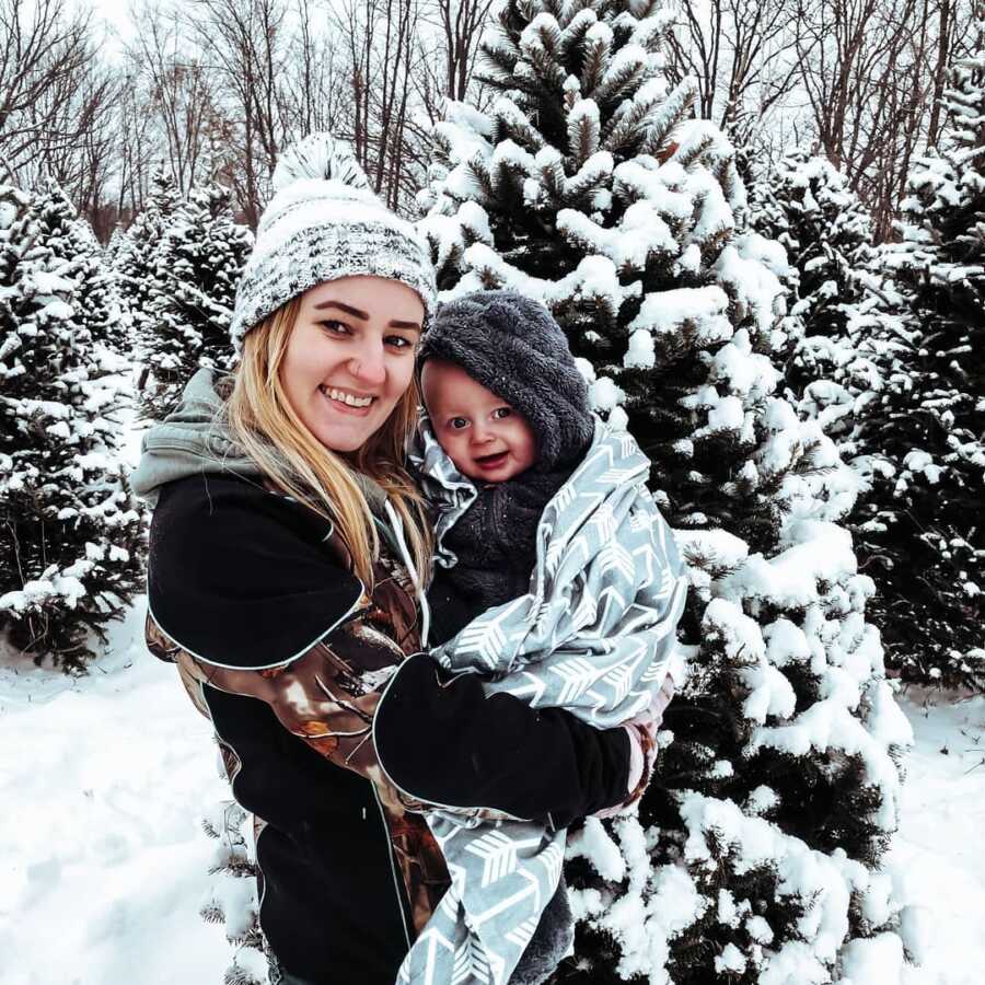 Young mom holds her baby boy, snuggled up in a warm blanket, outside in front of snow covered pine trees.