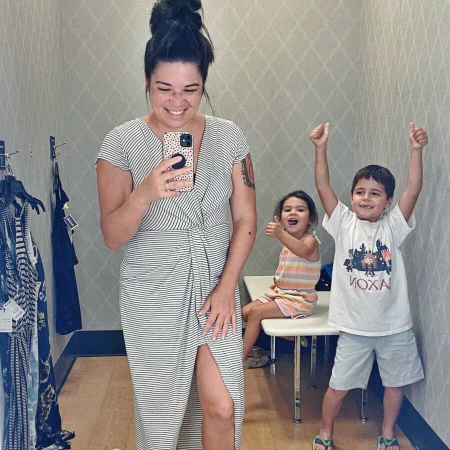 Mom trying on clothes in fitting room with children giving enthusiastic thumbs up in support. 