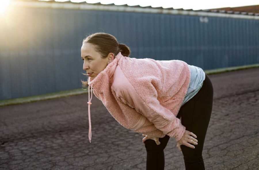 Woman exercising outdoors with her hands on her knees and leaning forward looking tired and wearing a pink sports jacket and black leggings with the sun in the back 