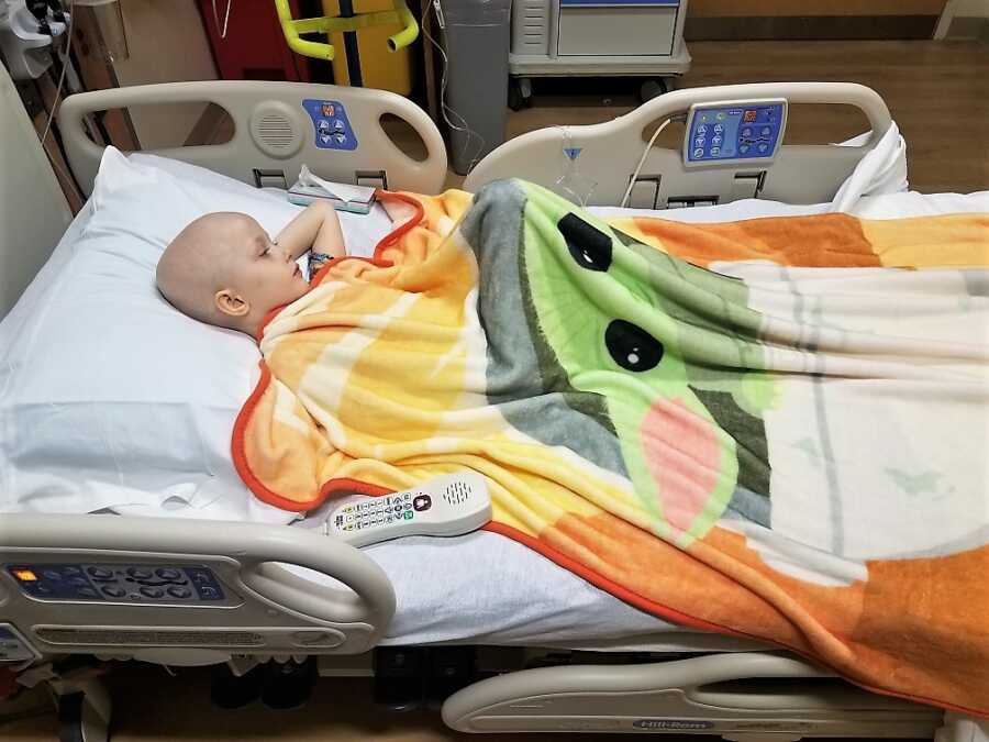 5-year-old kid diagnosed with Wilms Tumor laying on a hospital bed watching tv and covered with a baby Yoda blanket 