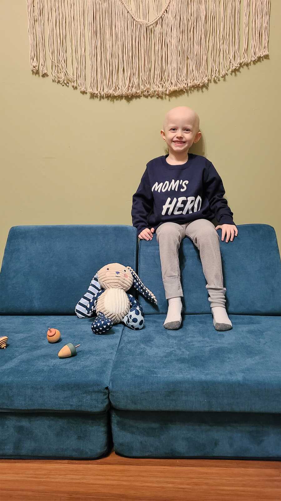 5-year-old cancer survivor sitting on a blue couch with a shirt that says 'Mom's Hero'