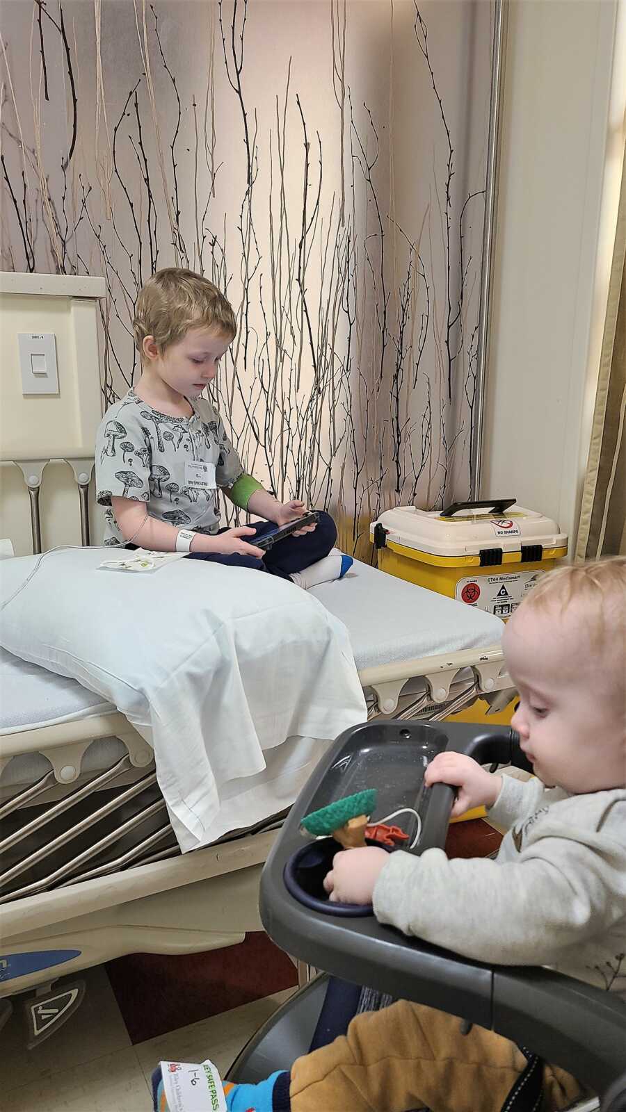 5-year-old kid diagnosed with Wilms Tumor receiving treatment, sitting in a hospital bed while watching videos on a phone next to his little brother 