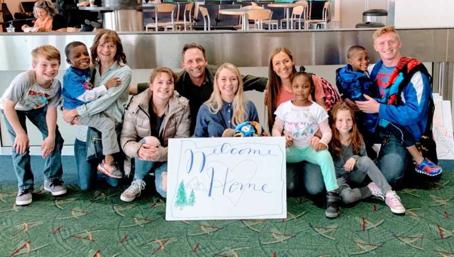 Family adopting three African American children smile big with a 'Welcome Home' sign at the airport