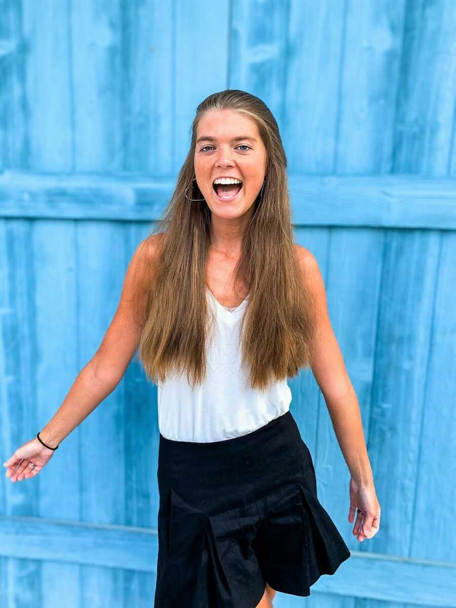 Young woman battling unknown chronic illness smiles big for a photo in front of a blue wall