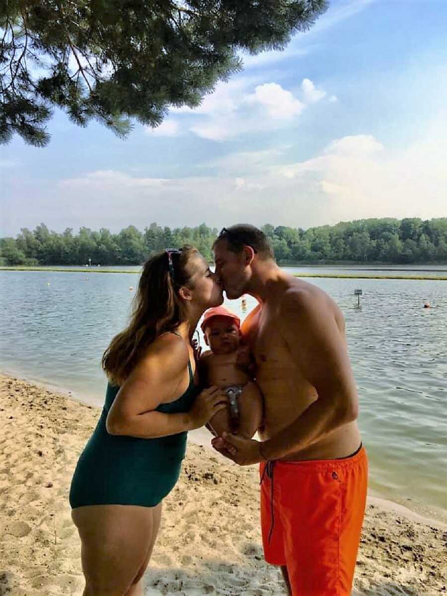 husband and wife kiss at the beach while holding son 