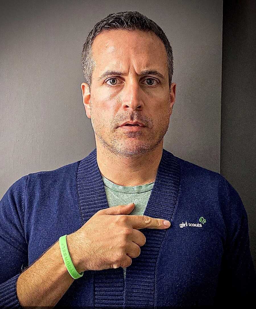 Father wearing blue Girl Scouts cardigan with a serious face and points to the Girl Scouts logo