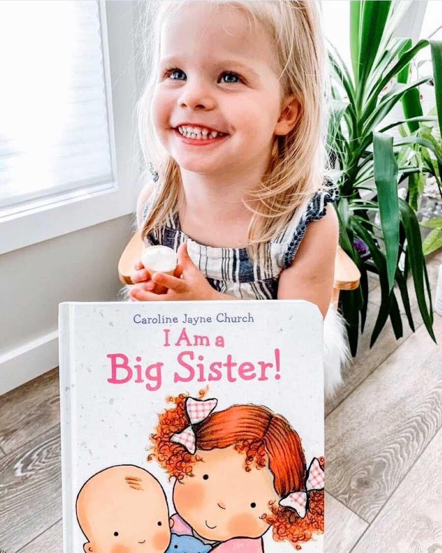 Little girl with blonde hair and blue eyes smiles while being gifted a book called "I Am a Big Sister!"