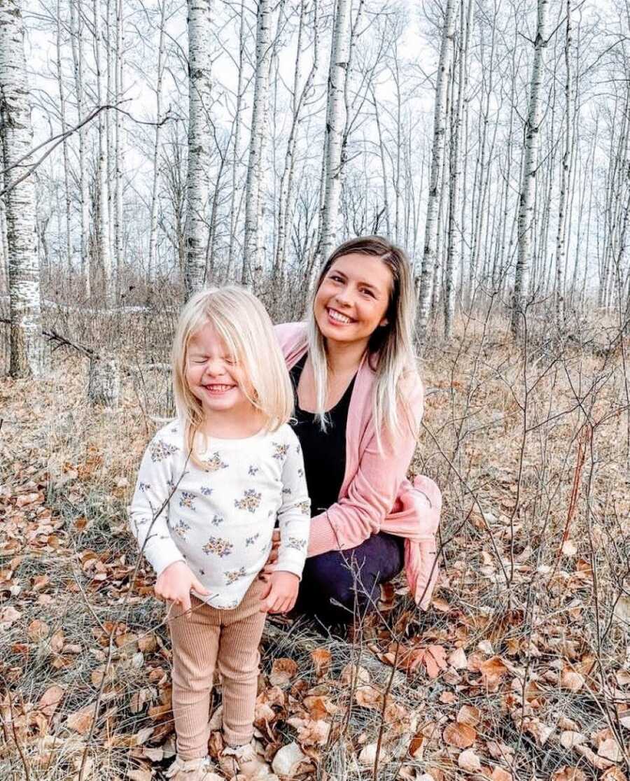 Pregnant mom squats down with her first daughter while on a walk in the woods