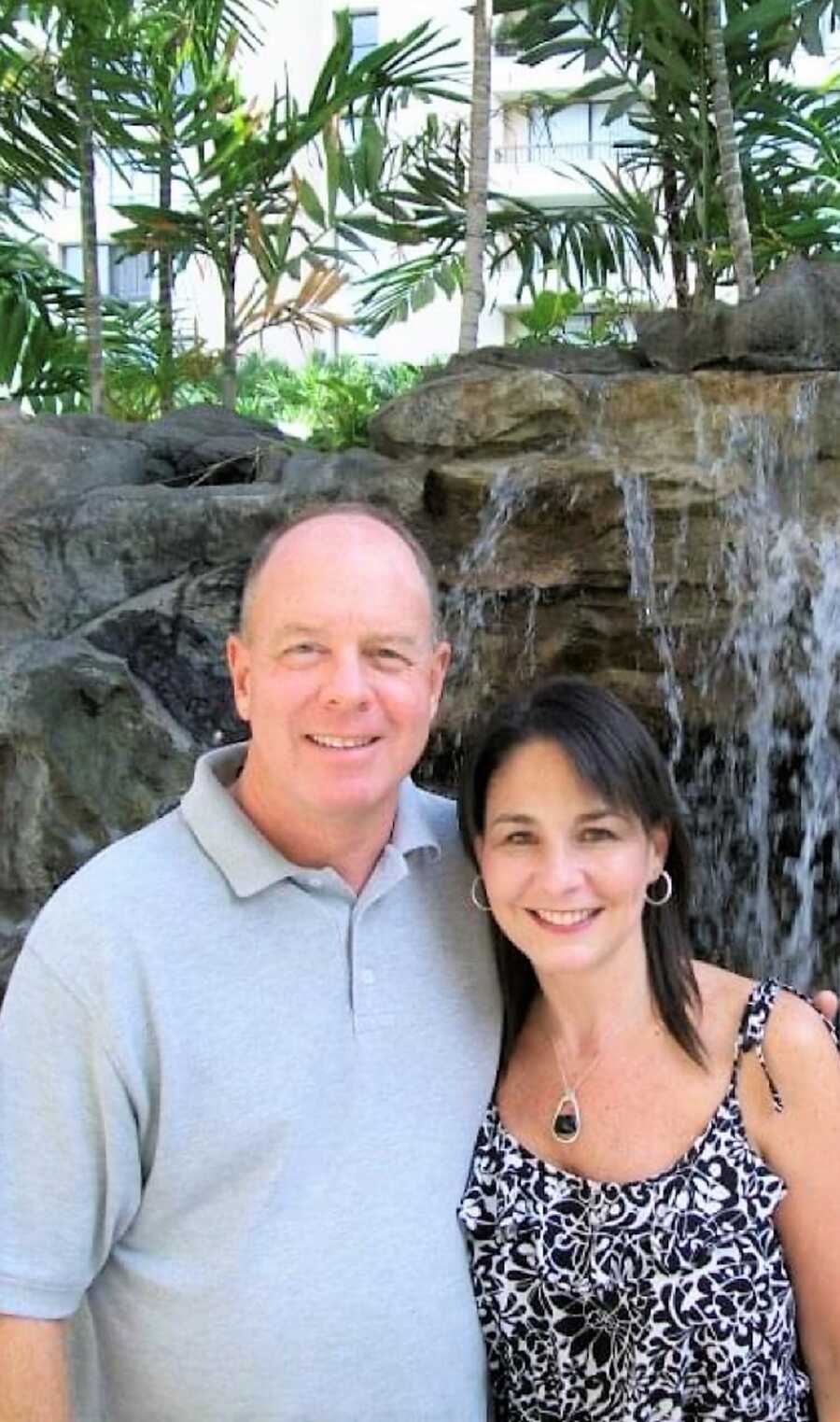 Happy husband and wife smiling in front of a waterfall with palm trees in the back