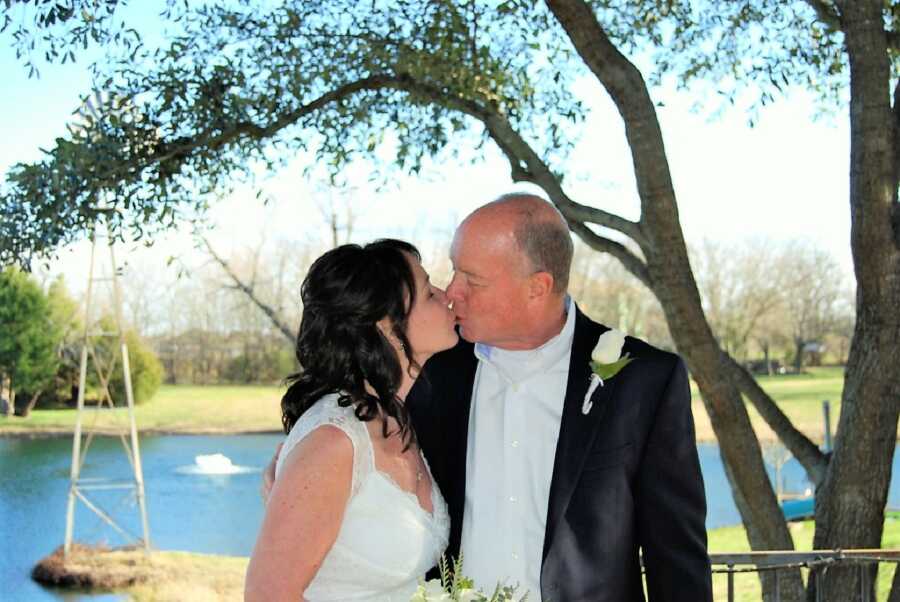 bride and groom kiss on their wedding day in front of a lake while holding a white rose bouquet 