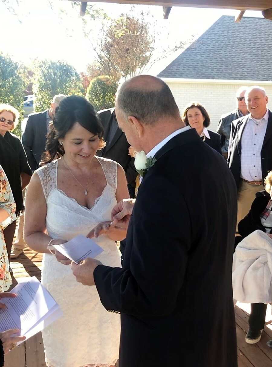 60 year old groom reading his wedding vows and putting the wedding ring on the bride's finger with the guests smiling in the back