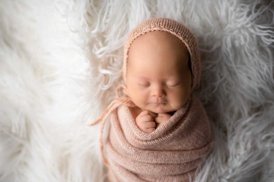 newborn baby photographed wearing a knitted outfit asleep on top of a white fuzzy blanket 