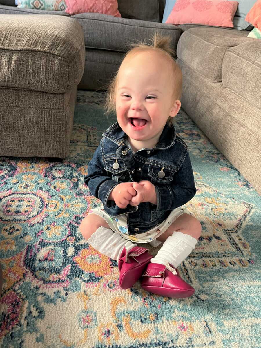 baby girl with down syndrome wearing red boots and denim jacket smiling big while sitting on the carpet in the living room 