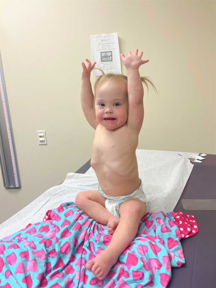 Down Syndrome baby celebrating with her arms up in the air sitting at the doctor's office 