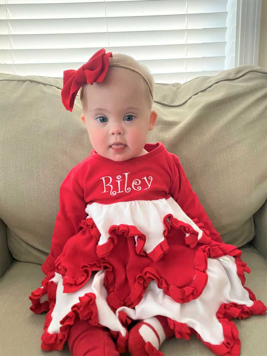 Baby girl with Down Syndrome sitting in the couch wearing a red and with dress with fringes and a big red bow on her head