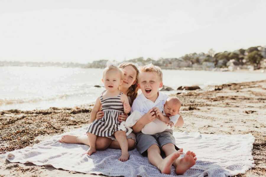 siblings sitting on a towel at the beach holding newborn baby and sister with down syndrome 