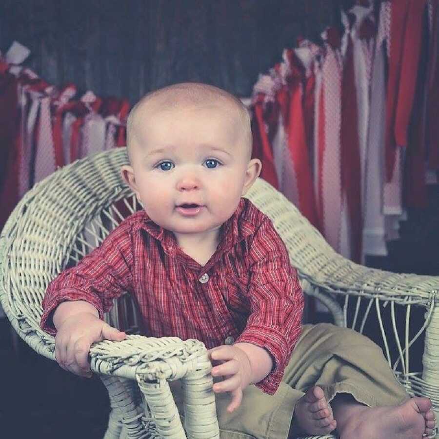 baby sitting in a chair wearing a red shirt and cargo pants with red ribbons hanging in the background 