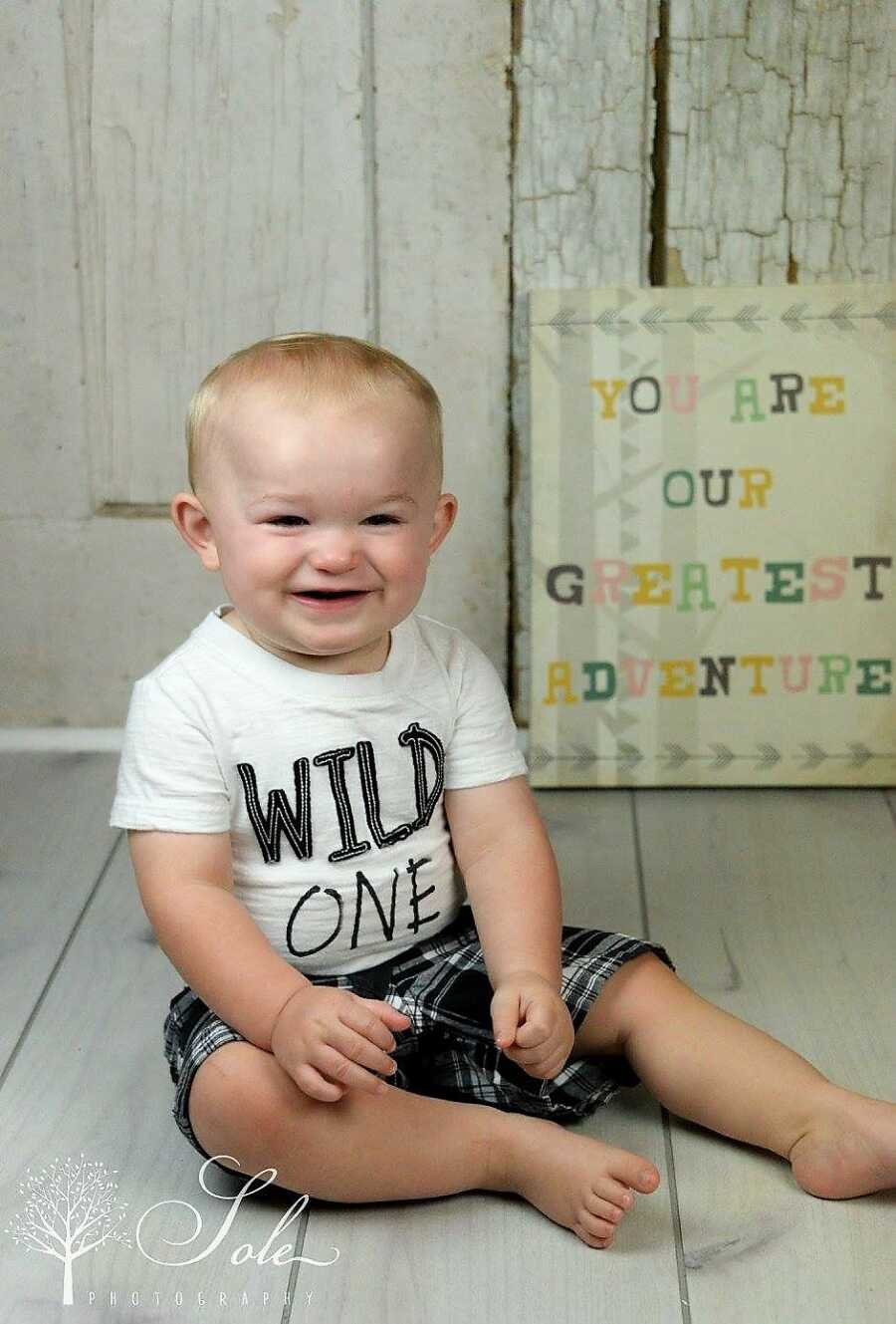 baby boy smiling wearing a white shirt that says 'Wild One' and a sing in the back that says 'you are our greatest adventure'