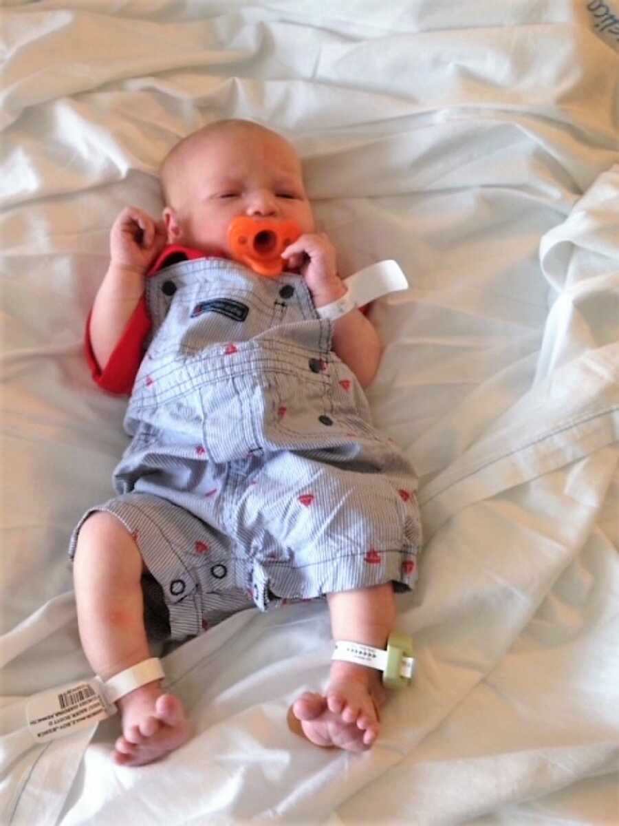 newborn baby wearing a denim overall with hearts and an orange pacifier laying on a bed with white sheets 