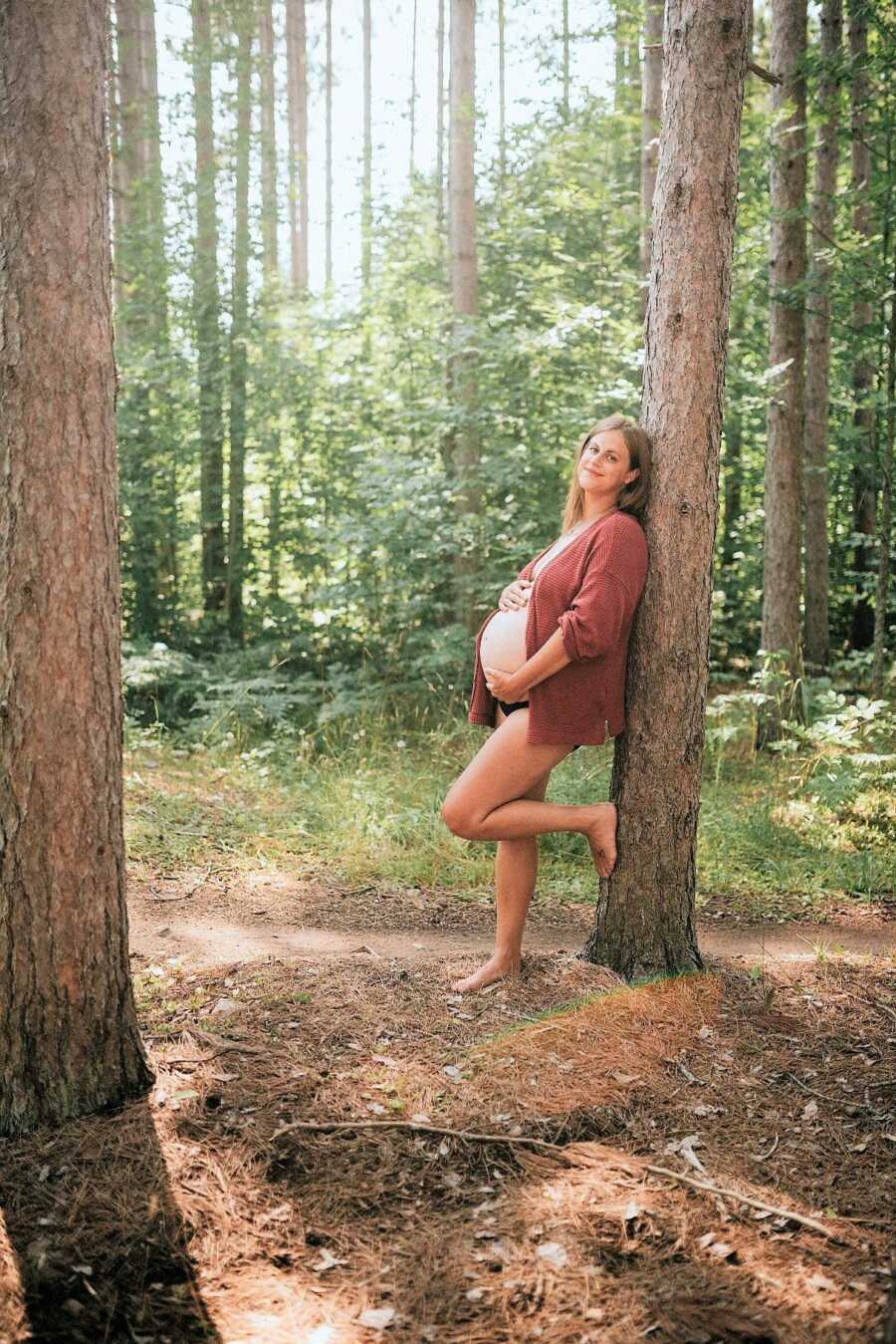 pregnant woman with barefoot resting over a tall pine tree holding her belly in the woods 