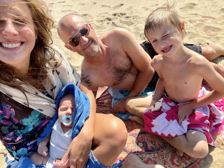mom, dad, son, and rainbow baby girl laying on the sand at the beach with the sun reflecting on their faces 