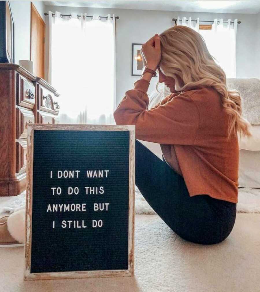 Woman struggling with infertility looks forlorn while sitting behind a sign that reads "I don't want to do this anymore but I still do"
