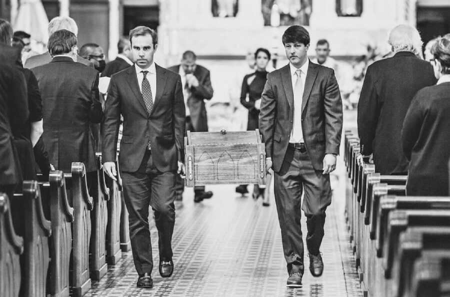 two man carrying casket during funeral with widow walking behind 