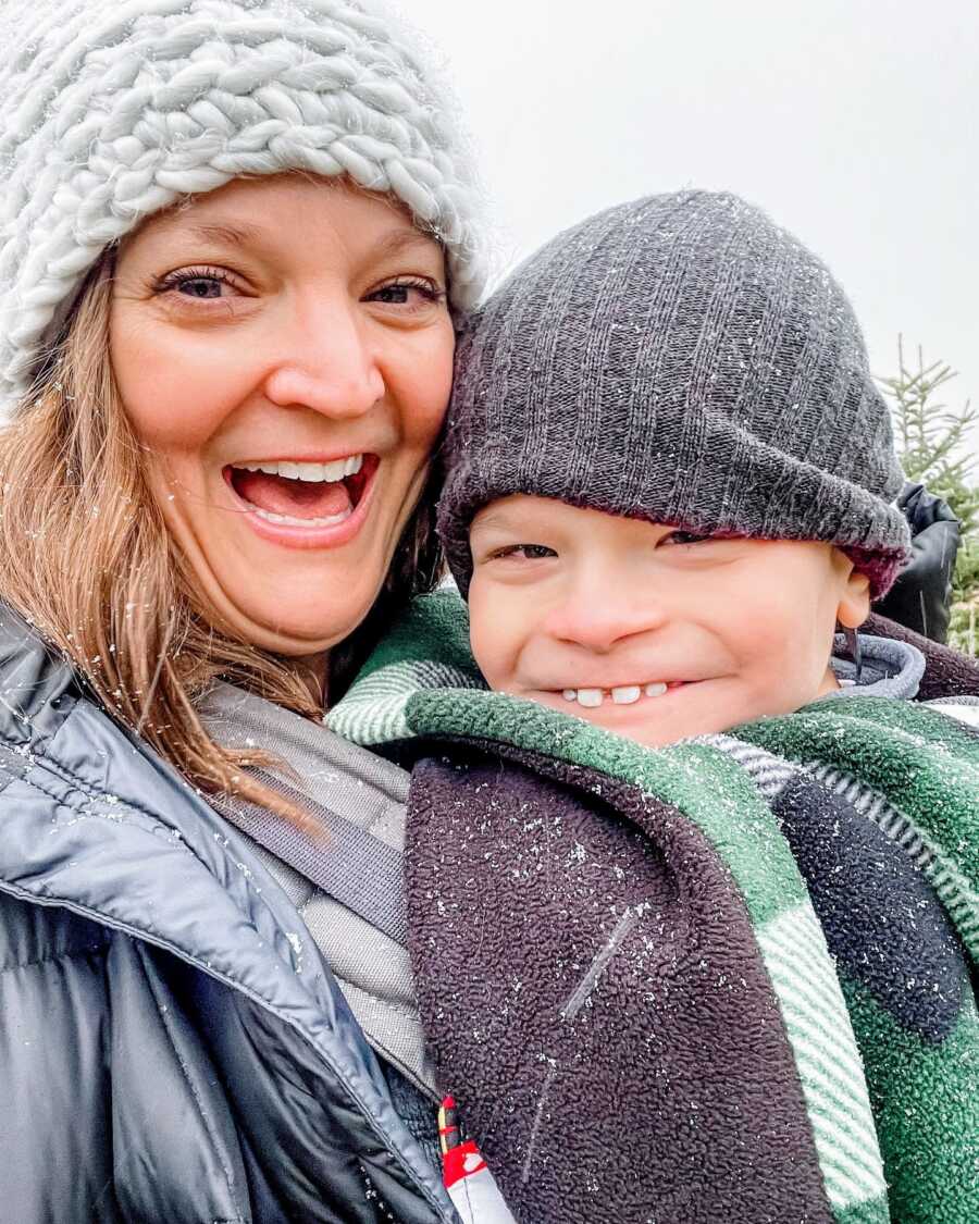 special needs mom carrying her son on her chest outdoors on a winter day while both wearing gray beanies and a blanket