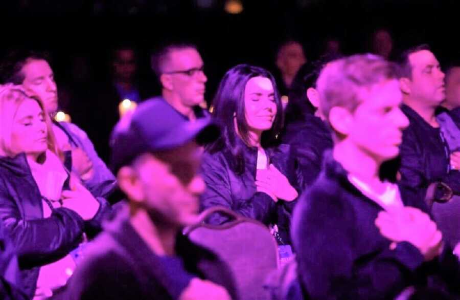 Empowerment coach for adult adoptees during a ceremony with the crowd placing the right hand on top of their hearts in purple lighting 